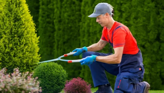 A professional gardener trimming a small shrub with long-handled shears, wearing a red shirt, blue pants, and gloves, set against a backdrop of tall conifer trees and vibrant flowers.