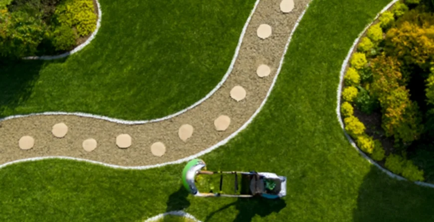 Aerial view of a gardener mowing a well-maintained lawn with a green mower, following a winding gravel path with round stepping stones, surrounded by vibrant plants and neatly trimmed grass.