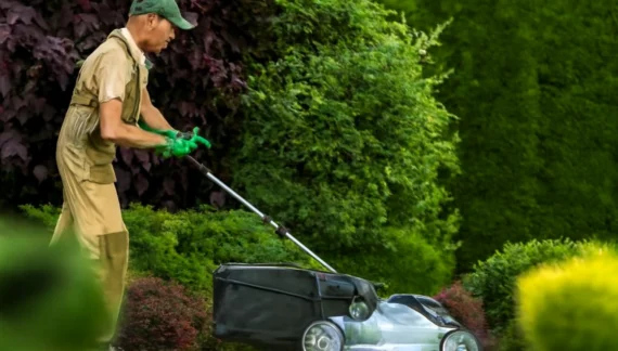 A landscaper operating a red zero-turn mower across a well-maintained lawn with neatly trimmed grass, set against a backdrop of tall trees and a white vinyl fence.